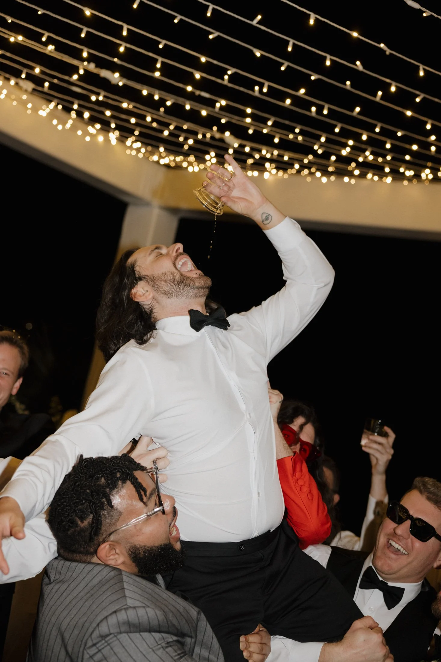 Group of men in formal attire celebrating at a party under string lights, lifting a groom holding a drink. Southern California wedding reception.