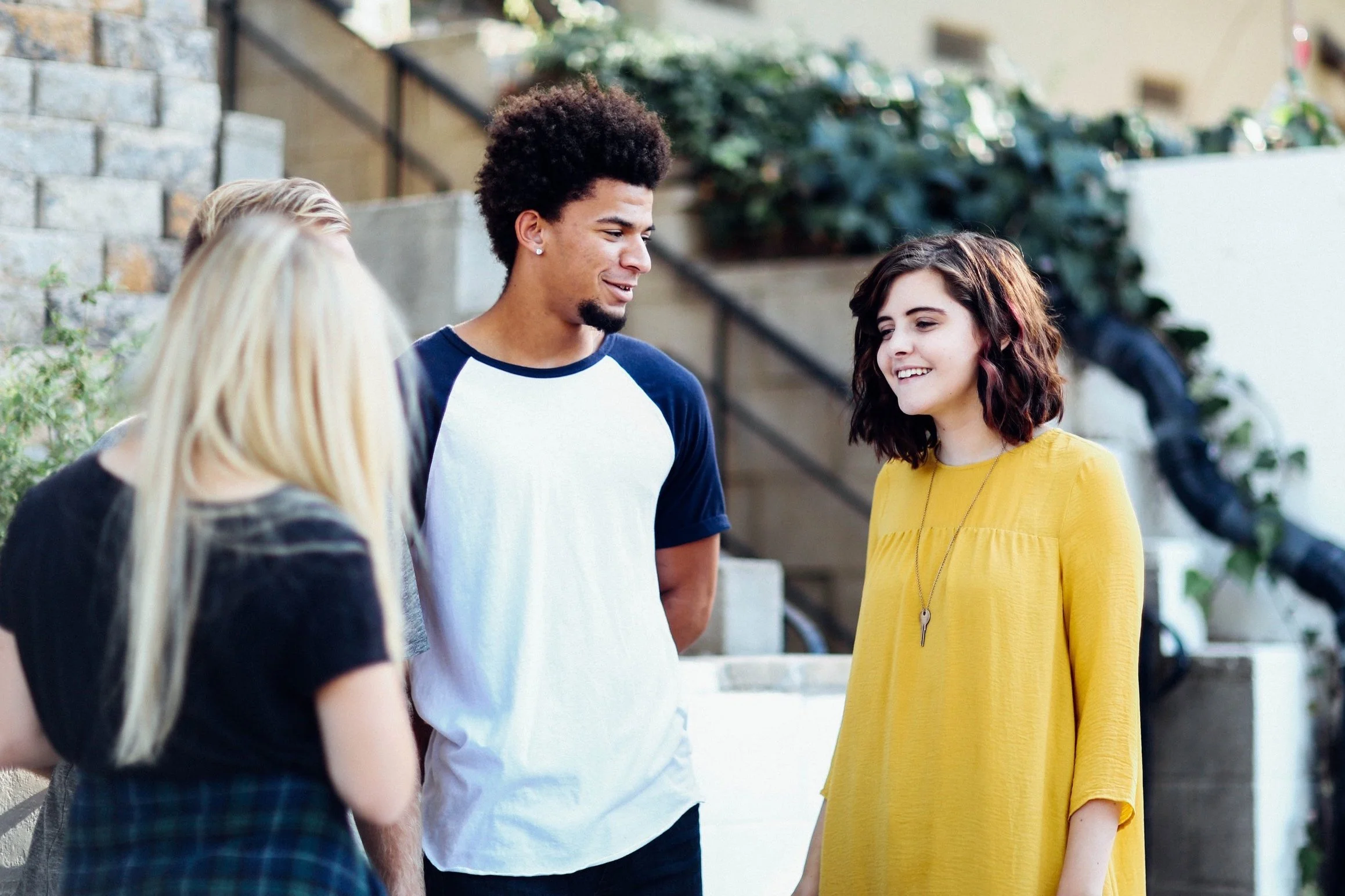 A group of four young adults standing outdoors, engaged in a conversation. One woman with blonde hair and a black top, another woman with wavy brown hair and a yellow dress, and a man with short curly hair and a white and blue baseball tee. They appear to be smiling and enjoying each other's company.