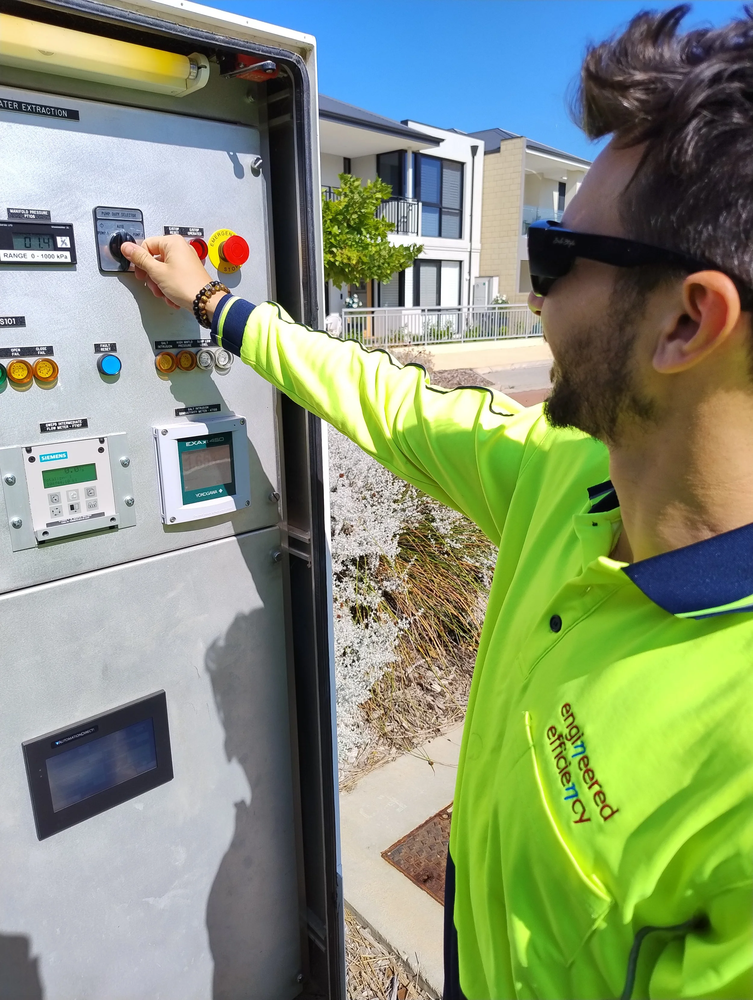 A man wearing sunglasses and a bright yellow high-visibility jacket operating a water extraction control panel outdoors on a sunny day with modern residential buildings in the background.