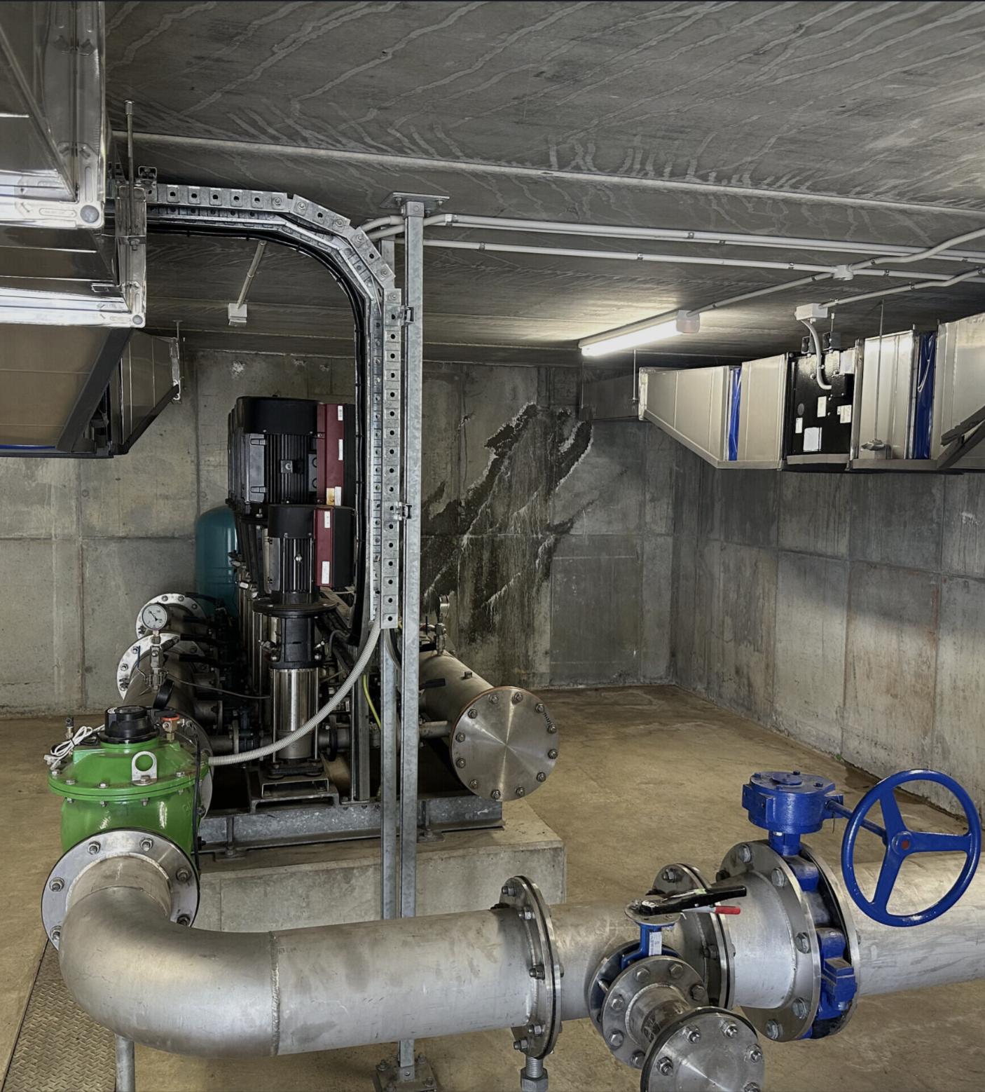 Industrial mechanical room with pipes, valves, and machinery, including a large horizontal pipe with a blue valve handle, a green pump, and electrical panels, against concrete walls and ceiling.