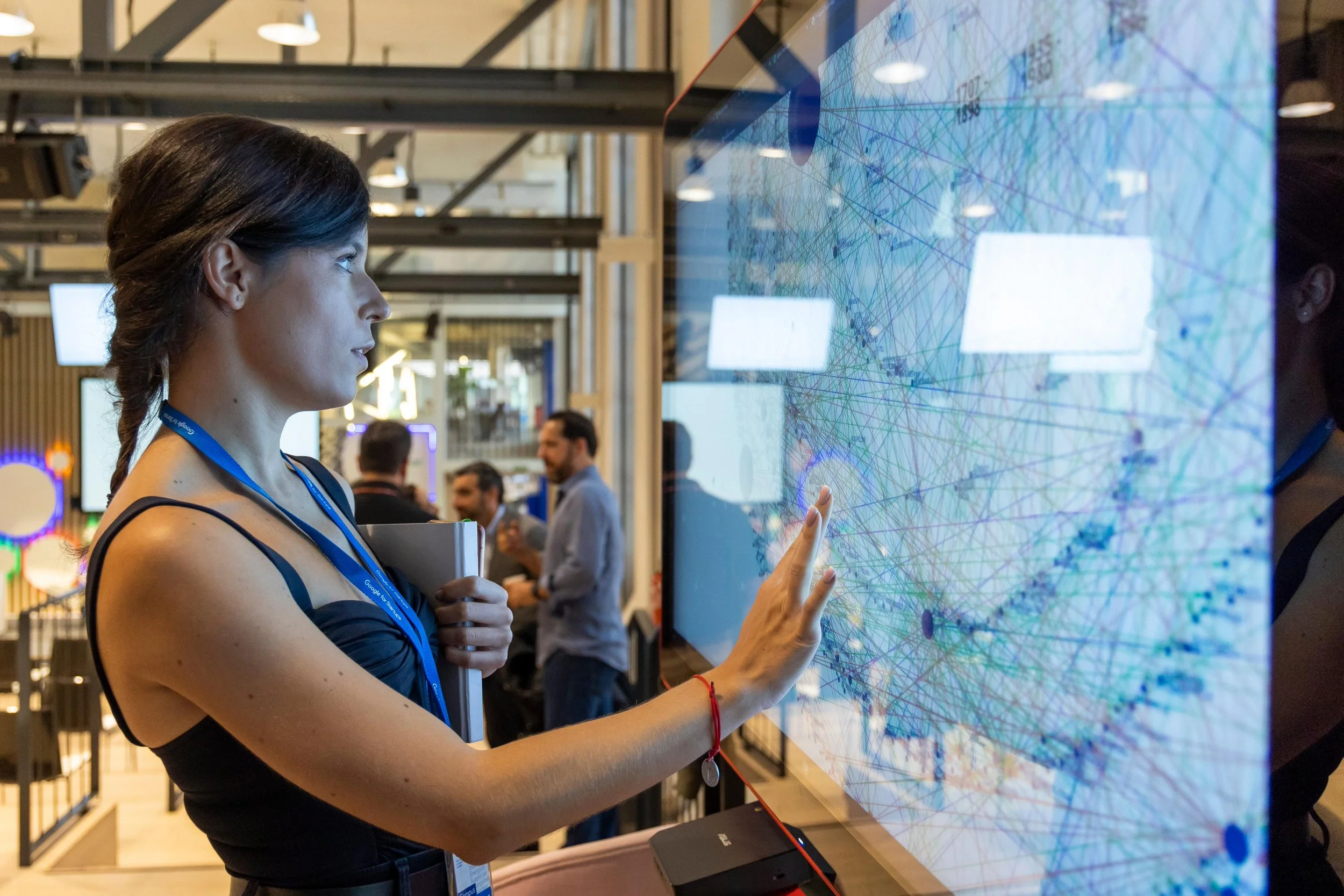 A woman interacting with a digital screen at a tech event, in a crowded space with several people in the background.