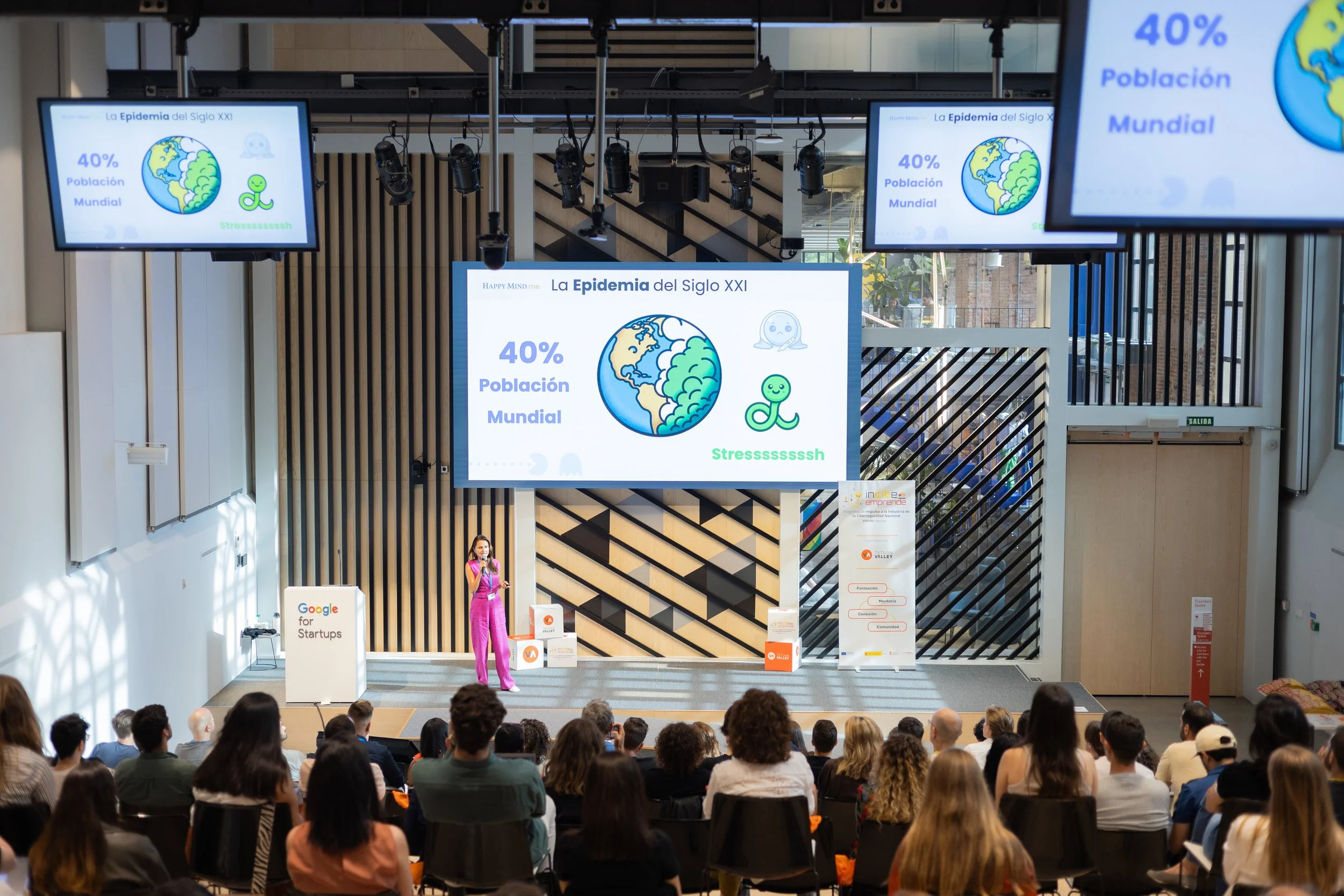 Conference in an auditorium with multiple screens showing a presentation titled 'The 21st Century Epidemic' with figures of 40% of the world population, and an illustration of planet Earth and a bacteria icon. Attendees are seated watching the talk, with a presenter standing on stage.