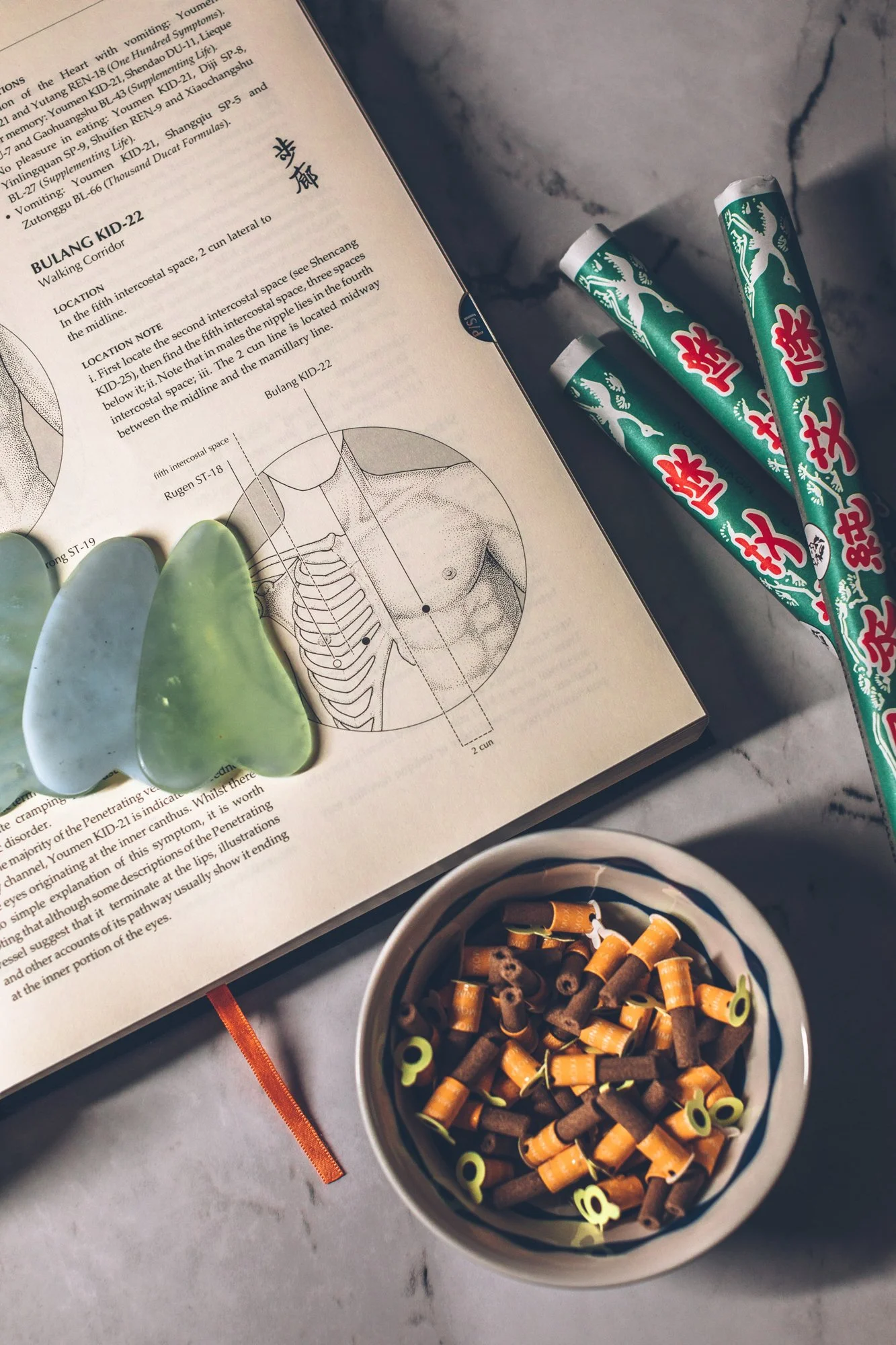 Jade gua sha tools and moxa rolls resting on an open acupuncture reference book.