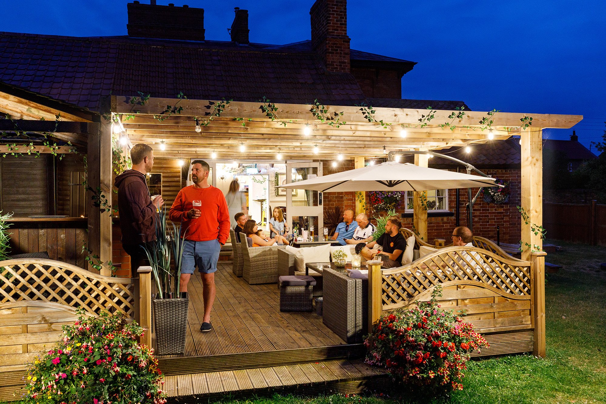 People socializing on a well-lit outdoor wooden patio with furniture and a large white umbrella, attached to a house during evening.
