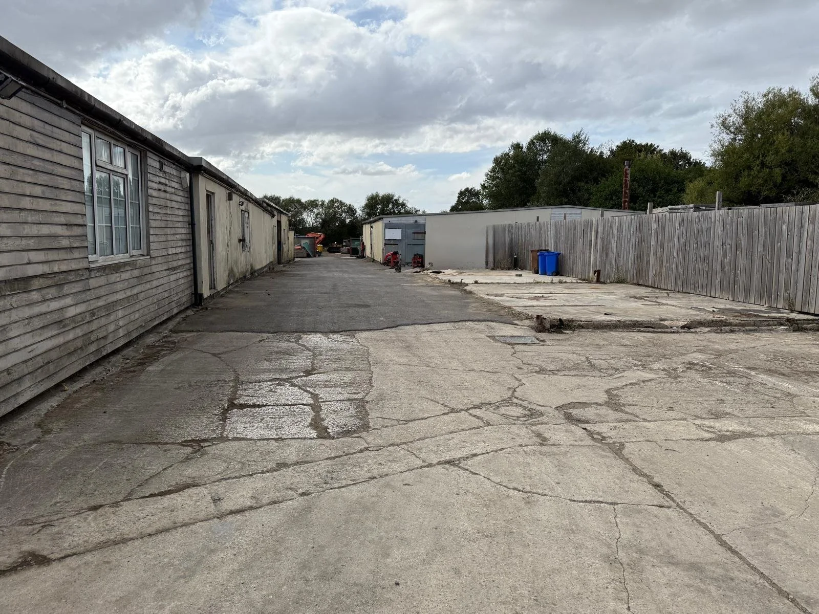 An alleyway between two buildings with a weathered wooden fence to the right and a building with siding to the left. The ground is cracked and uneven, with some patches of asphalt. There are a few trash bins and small objects further down the alley, with trees and a cloudy sky overhead.