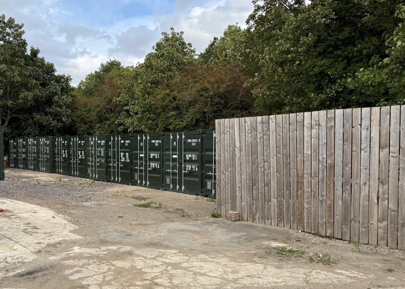 Line of green shipping containers next to a wooden fence in an outdoor area with trees and a cloudy sky in the background.