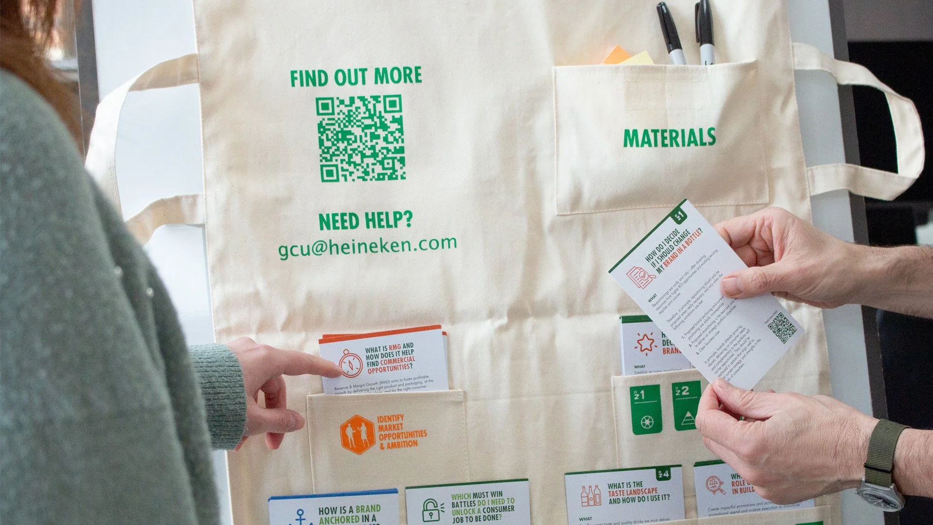 Two people holding informational cards in front of a beige fabric bag with green text and QR codes, labeled 'Materials' and 'Find Out More'.
