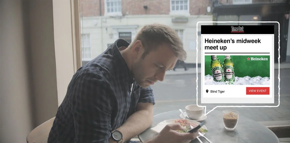Man sitting at a table in a cafe looking at his phone with a virtual overlay showing a Heineken beer event invite.