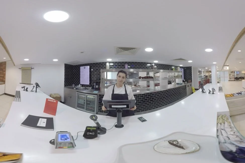 Reception area at a restaurant or cafe with a female staff member standing at the counter, surrounded by a cash register, payment terminal, and a plate with a slice of cake.