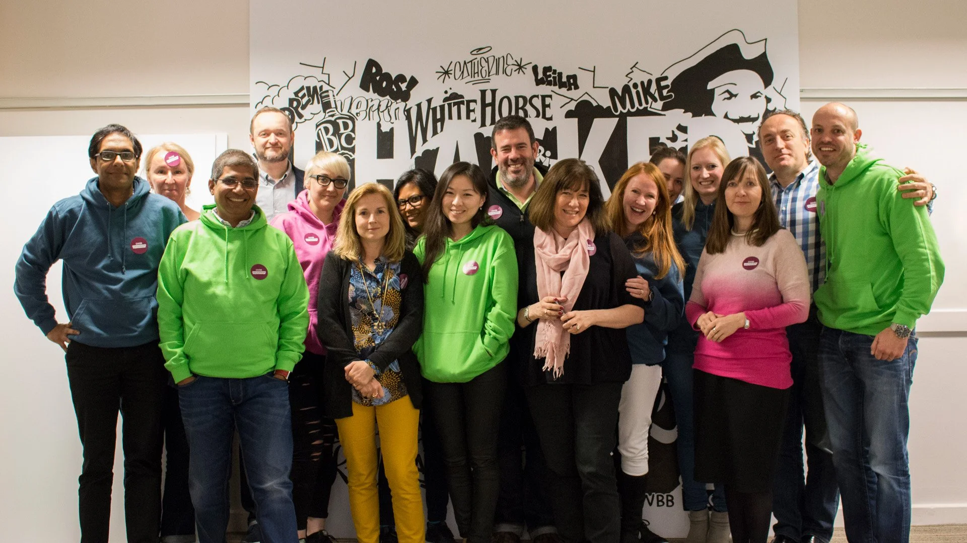 Group of 14 diverse people posing for a photo in front of a wall with text and illustrations, some wearing colorful jackets and badges.