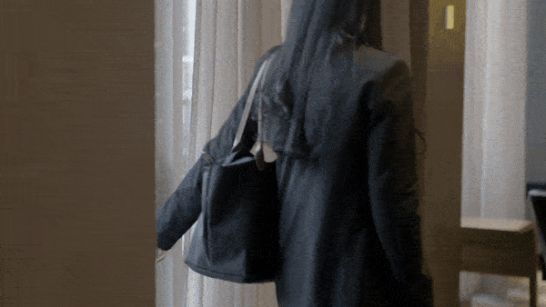 A woman with long dark hair wearing a black leather jacket carrying a black shopping bag, walking inside a building near a wall with a window.