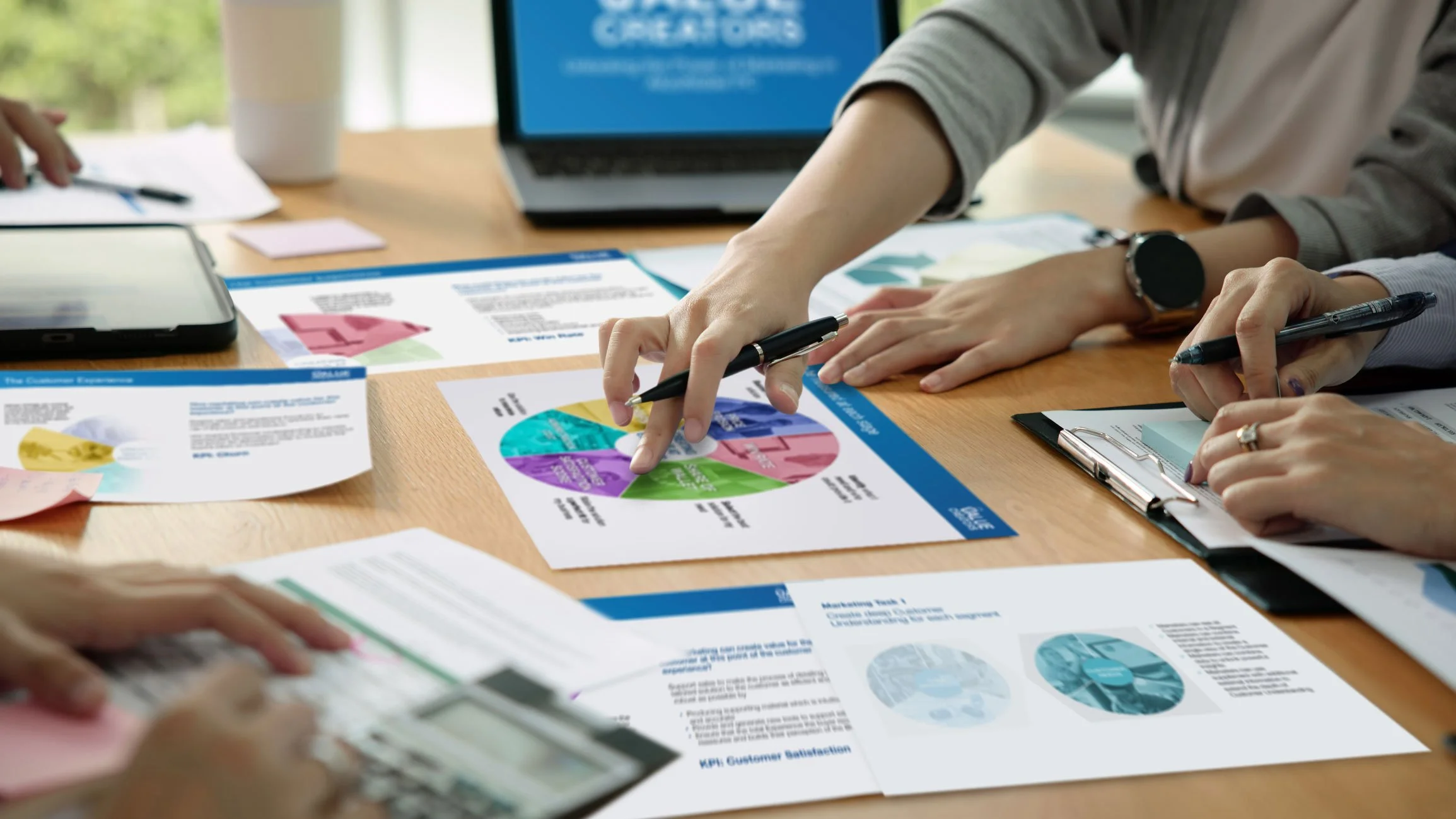 People working at a wooden table with documents, graphs, and charts, pointing at a colorful pie chart in a business meeting.