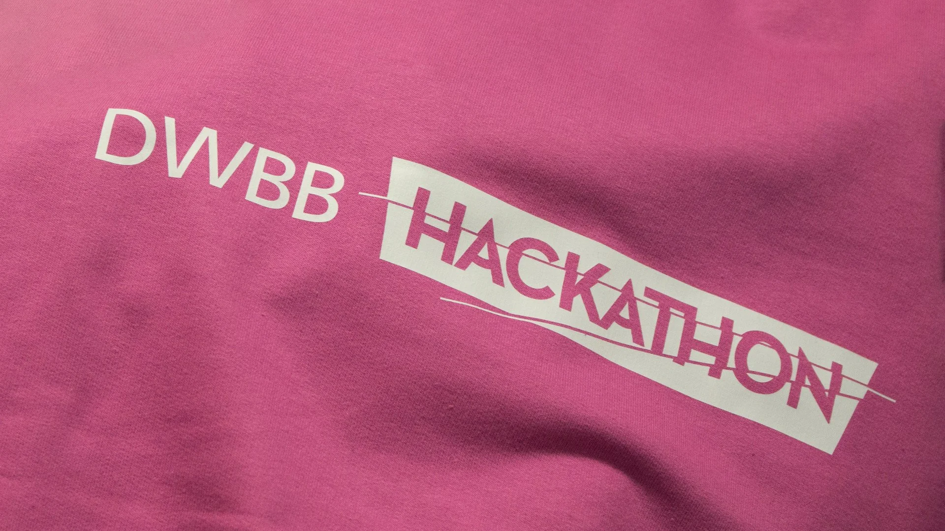 Pink T-shirt with white and purple text reading "DWB Hackathon" in a stylized, wavy font.