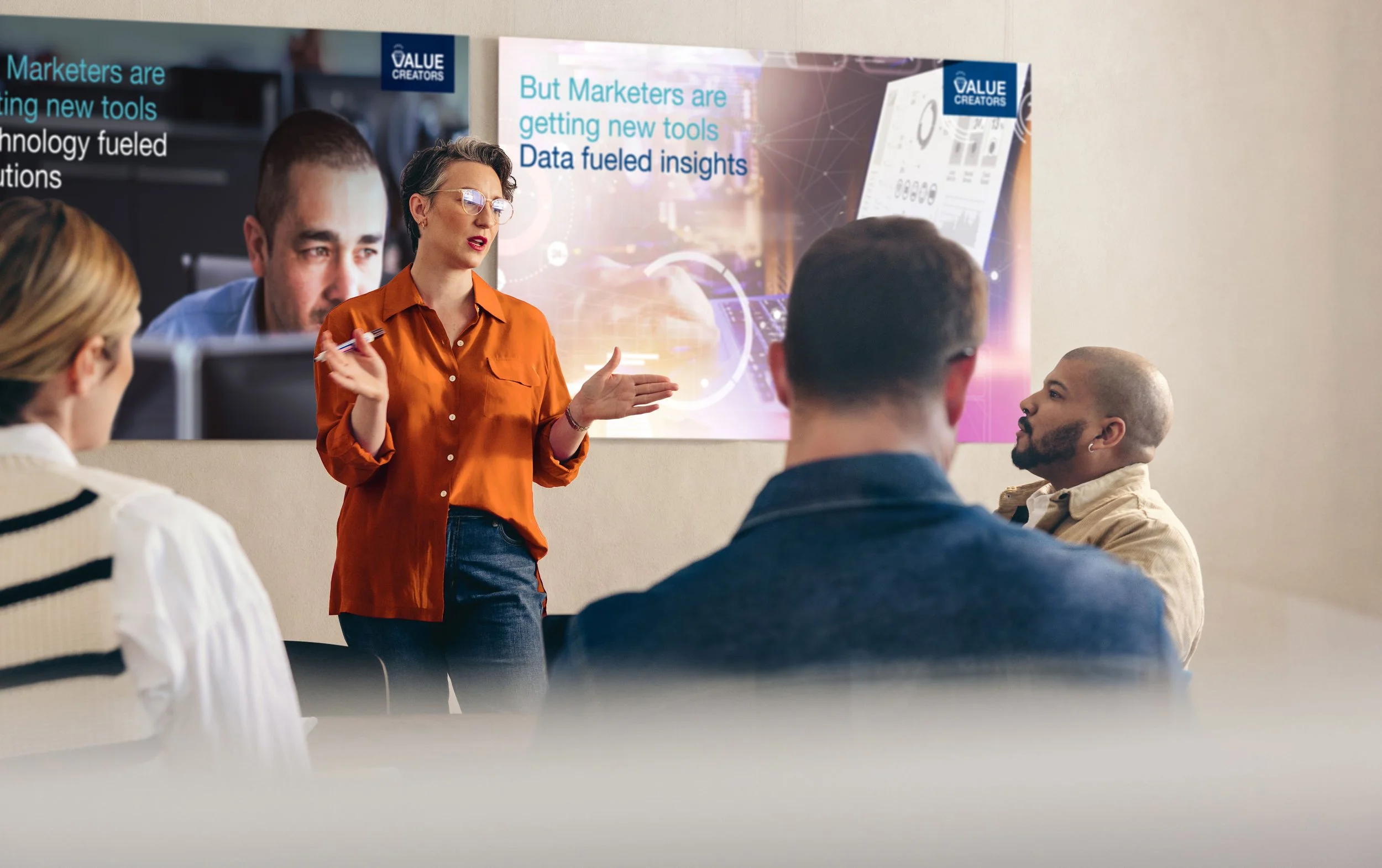A woman in an orange shirt giving a presentation to a group of four people in a room with a banner that reads, 'But Marketers are getting new tools, Data fueled insights.'