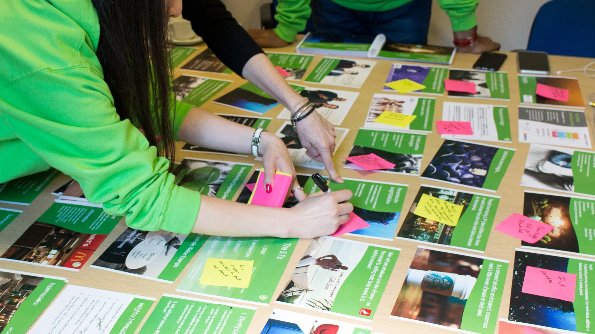 People gathered around a table reviewing green leaflets, some with pink and yellow sticky notes attached, using pens to make notes or mark information.