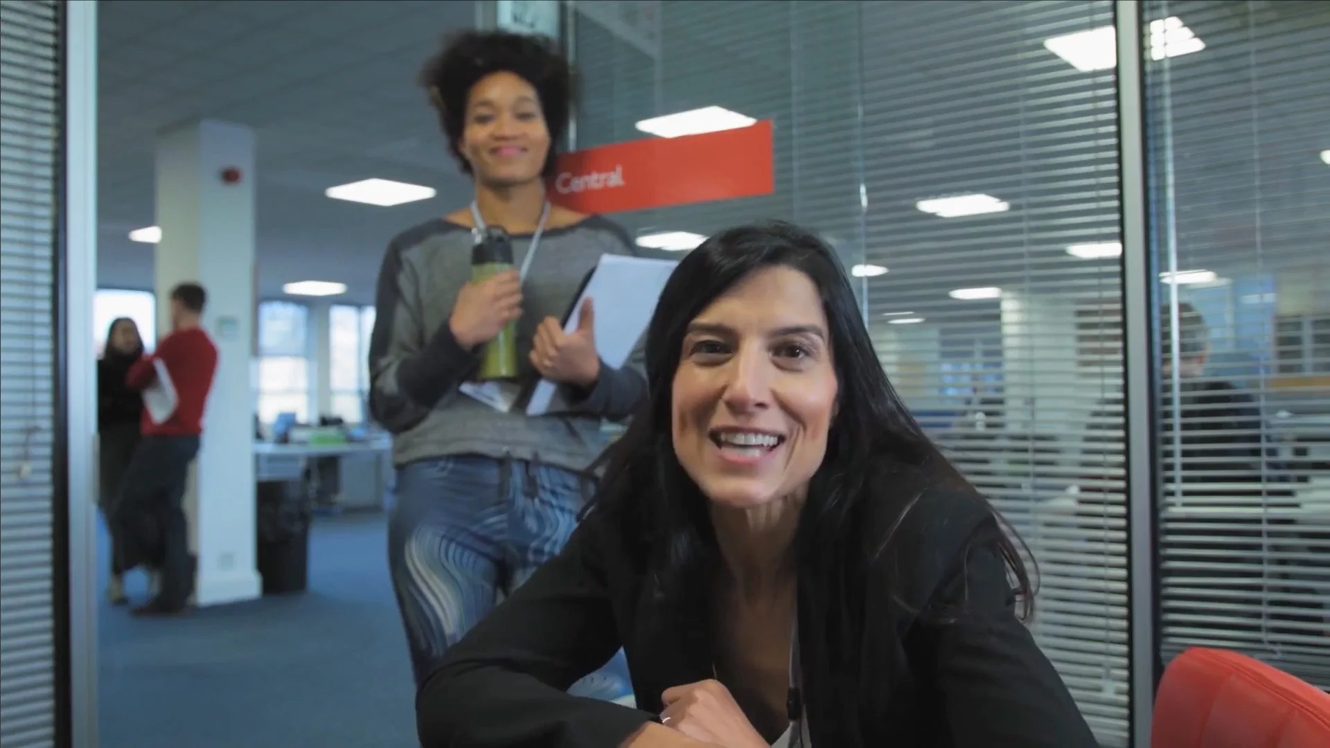 A woman with black hair smiling at the camera in an office environment, with a person standing behind her holding a bottle and notepad, and other people in the background.
