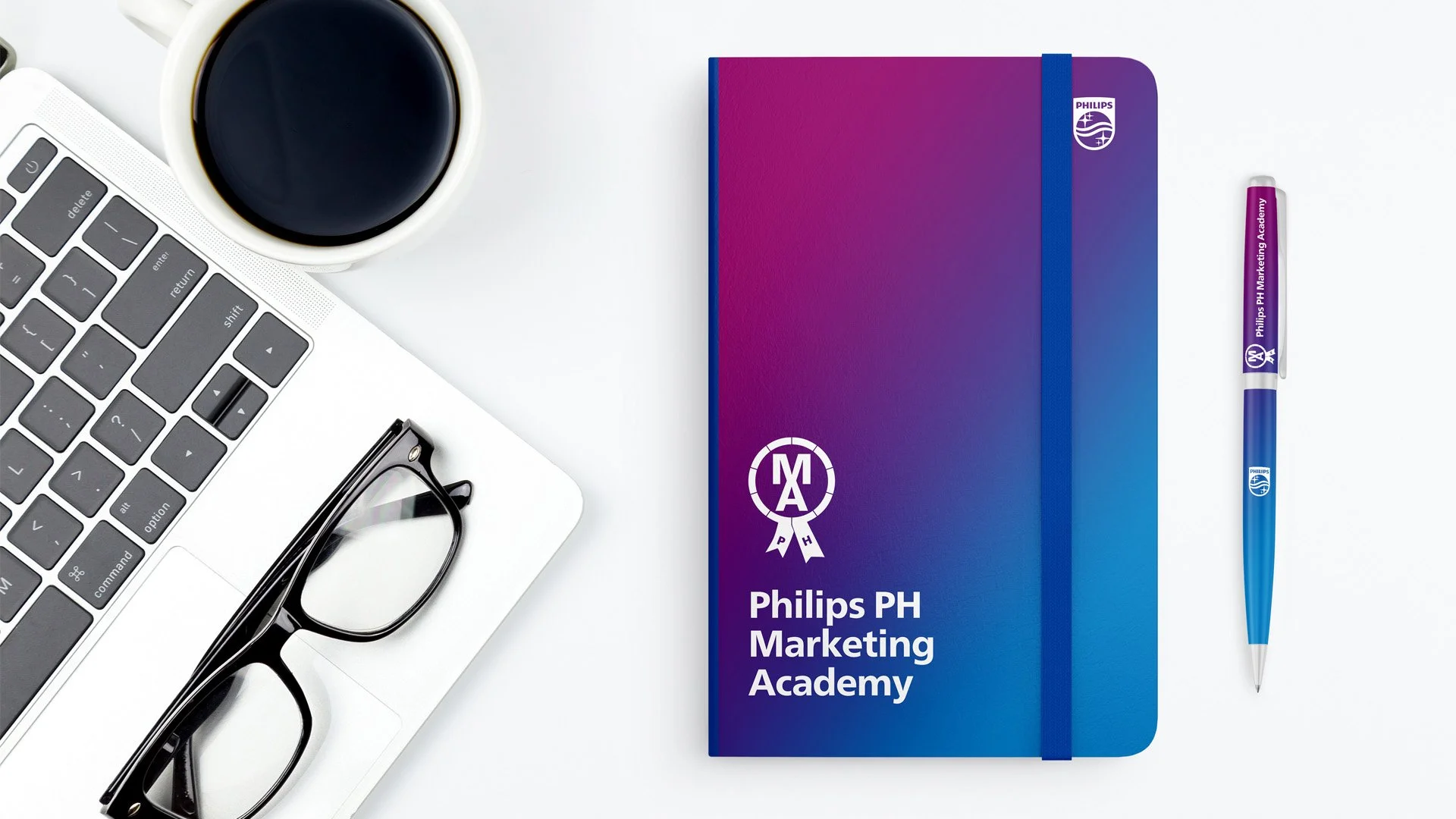 A workspace featuring a laptop with glasses on it, a cup of coffee, a purple and blue notebook labeled 'Philips PH Marketing Academy', and a matching pen on a white surface.