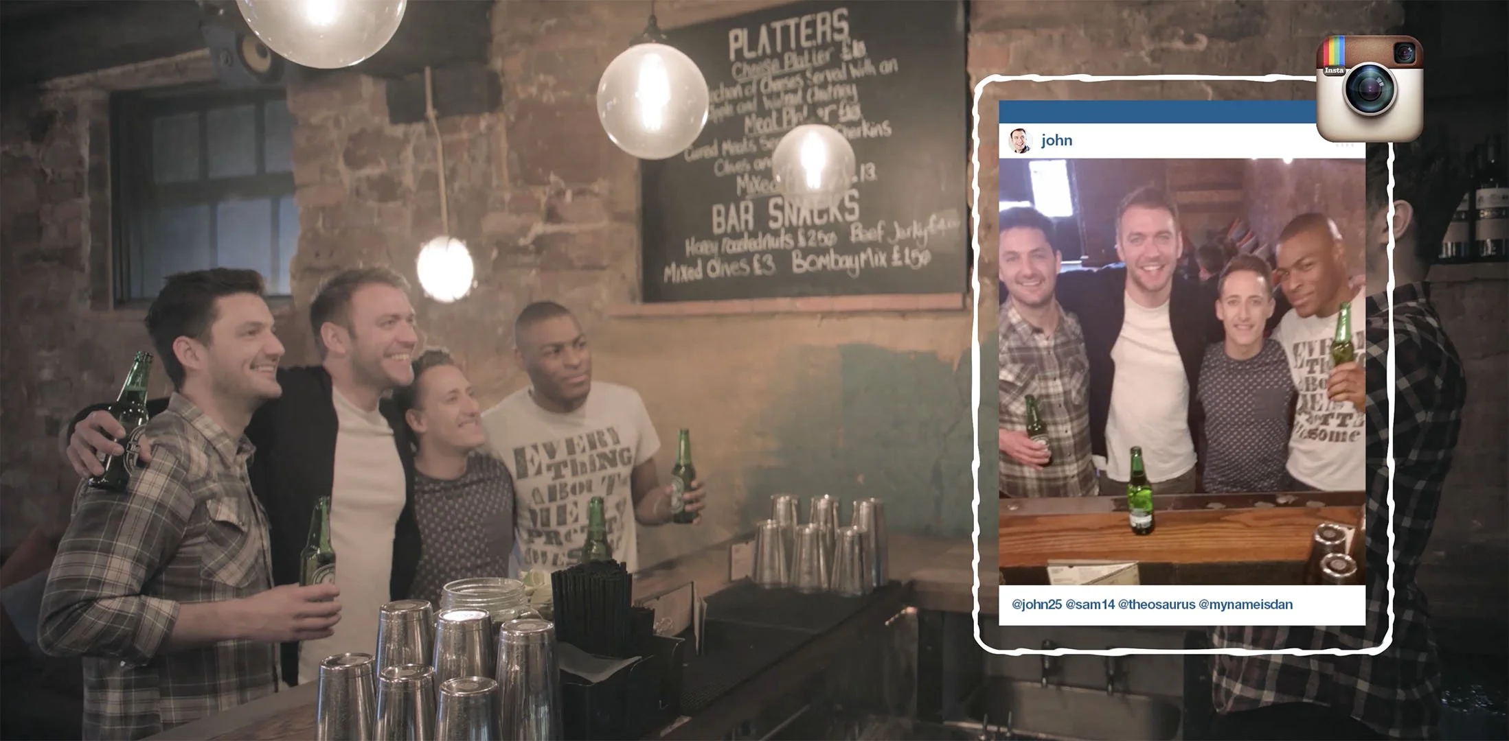 Group of friends at a bar taking a photo, with four men holding drinks and smiling, and a woman standing in the middle. The bar has a rustic brick wall with a chalkboard menu, and there are empty glasses on the counter. The photo is framed with an ov
