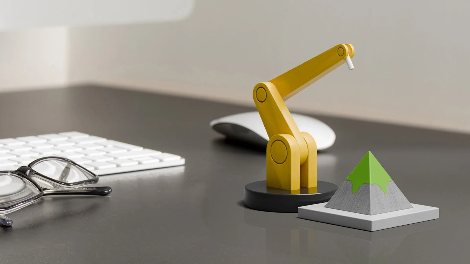 Desk with a pair of glasses, a white keyboard, a white computer mouse, a yellow wooden robot model, and a small chalkboard shaped like a mountain with green and gray coloring.