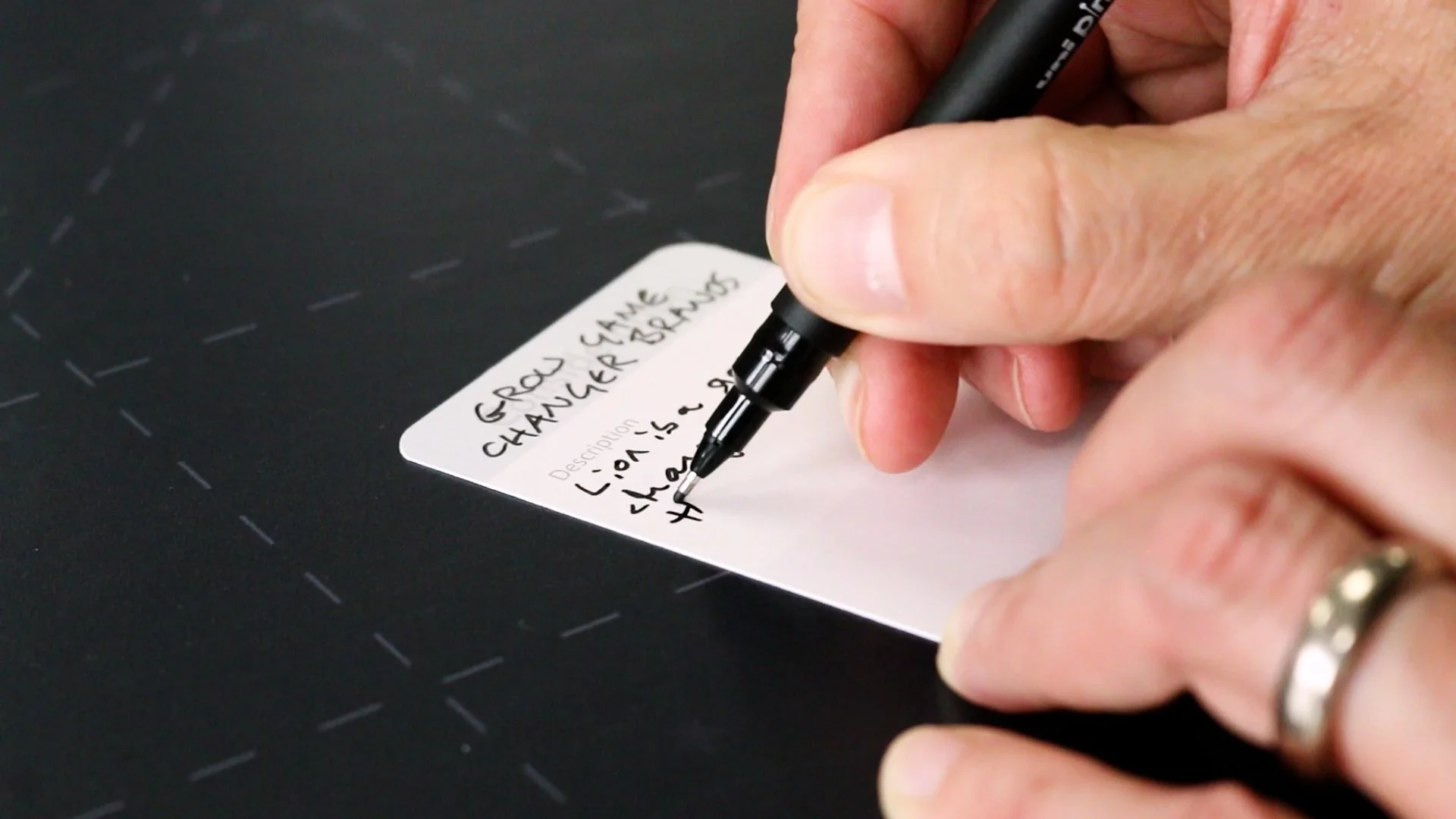 A person's hand writing on a white label with a black marker, with the label placed on a black surface with faint white dashed lines.