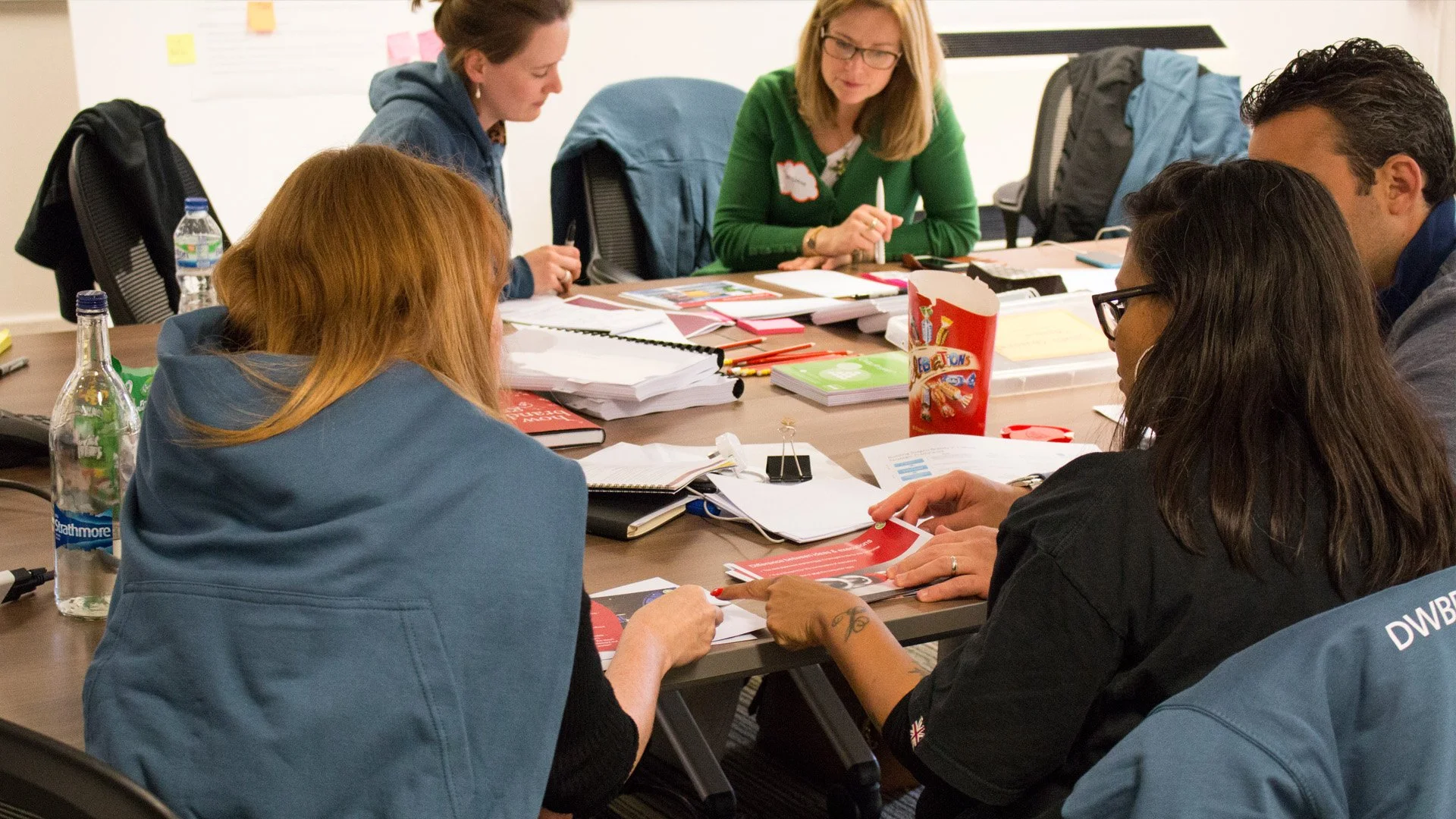 A group of five people gathered around a conference table in a meeting or workshop setting, discussing documents and notes.