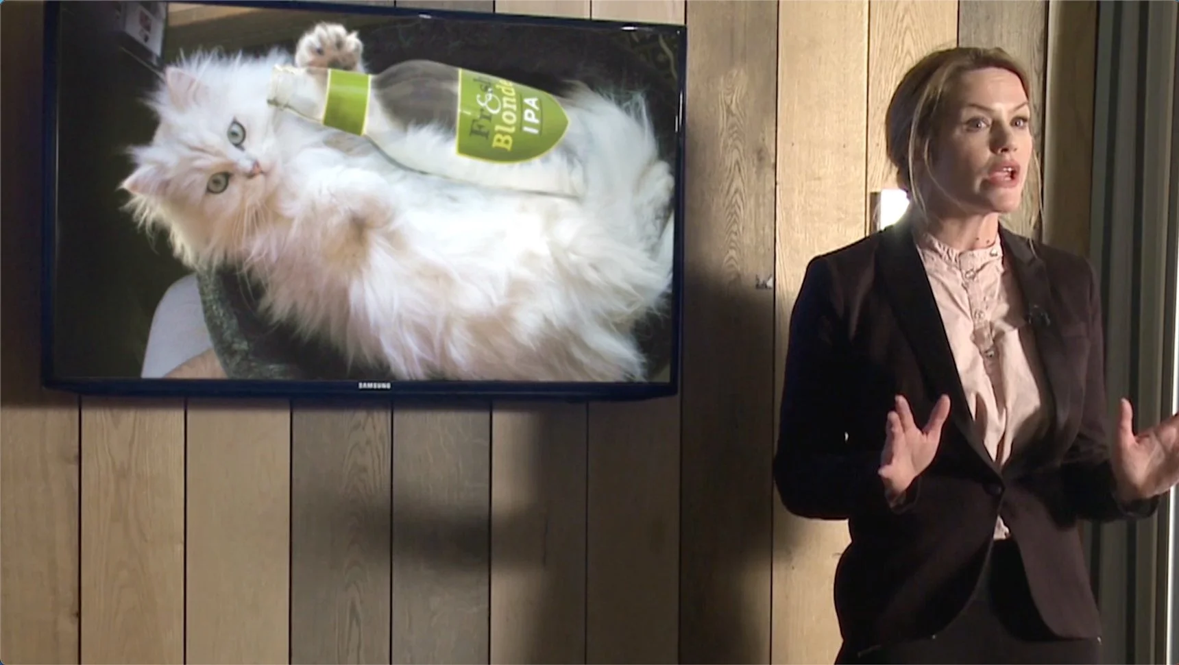 A woman is giving a presentation next to a large screen showing a cat with a bottle labeled "Tig & Bloom IPA." The cat is lying on its back with a bottle of beer on its belly.