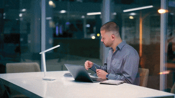 A man sitting at a desk in a modern office, looking at his smartphone beside a laptop and notepad, with large windows and city lights visible outside at night.