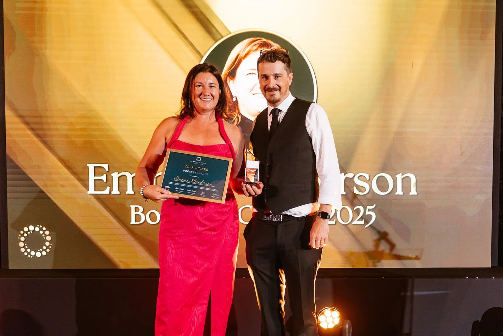 A woman in a pink dress holding an award plaque stands next to a man in formal attire. They are on stage at an event with a large screen behind them displaying a photo and the text 'Emma Henderson Bookers Choice 2025.' The woman is smiling and the man is holding a glass of wine.