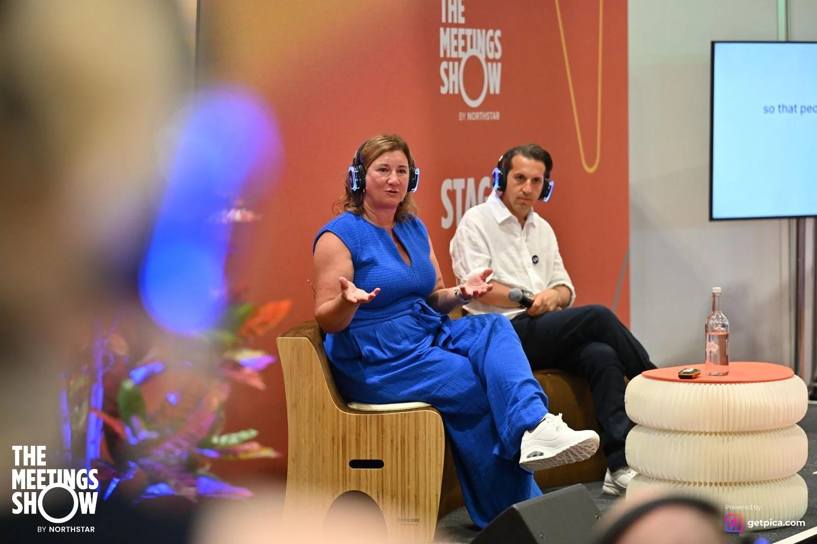 A woman and a man are sitting on stage at The Meetings Show, speaking into microphones while wearing headphones. The woman is wearing a blue dress and white sneakers, and the man is wearing a white shirt and black pants. A large screen is visible next to them, and a water bottle and a small device are on a round table in front of them.