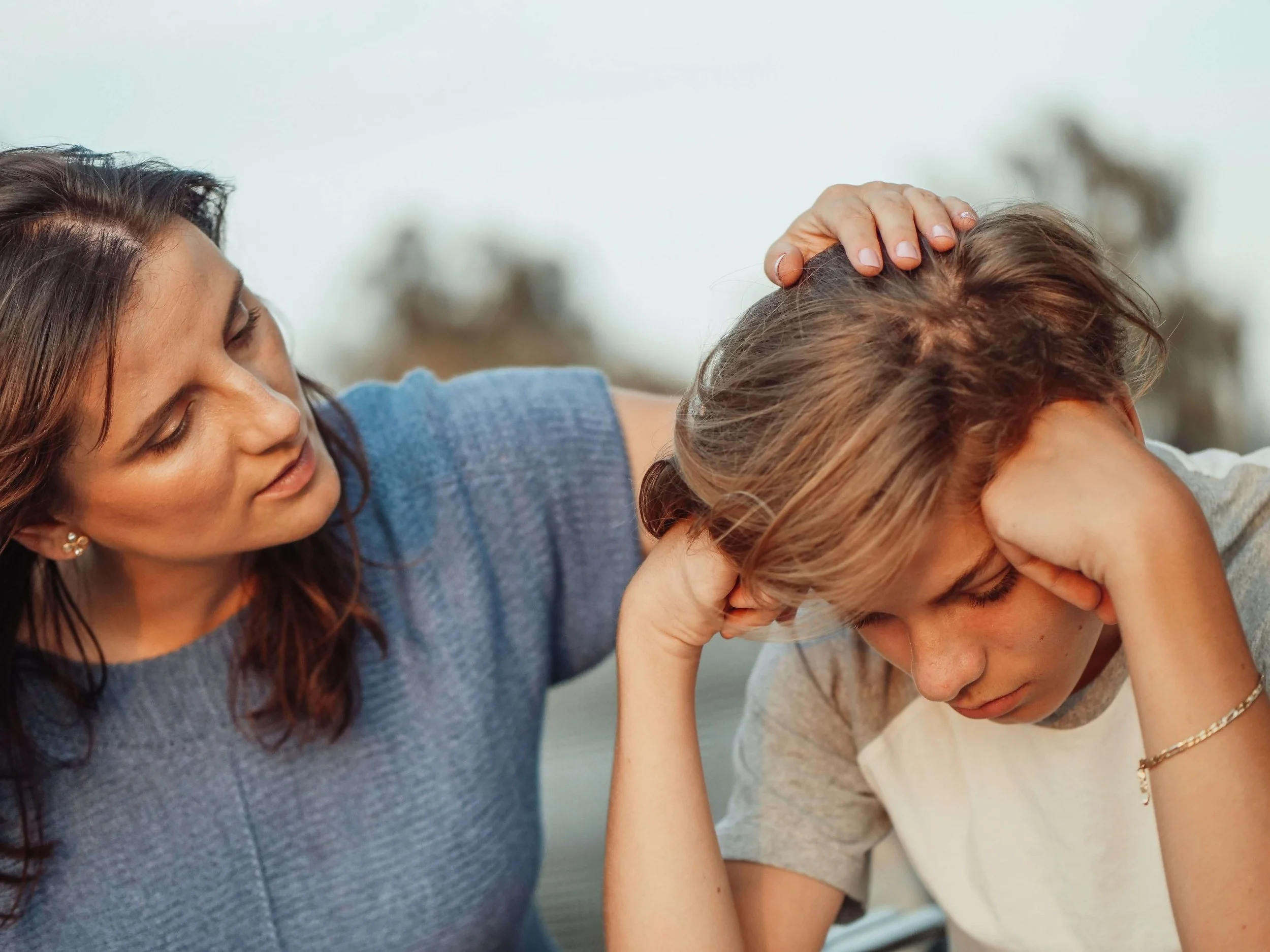 A woman with brown hair and a blue shirt comforting a young person with brown hair and a white shirt, who appears distressed, in an emotional moment.