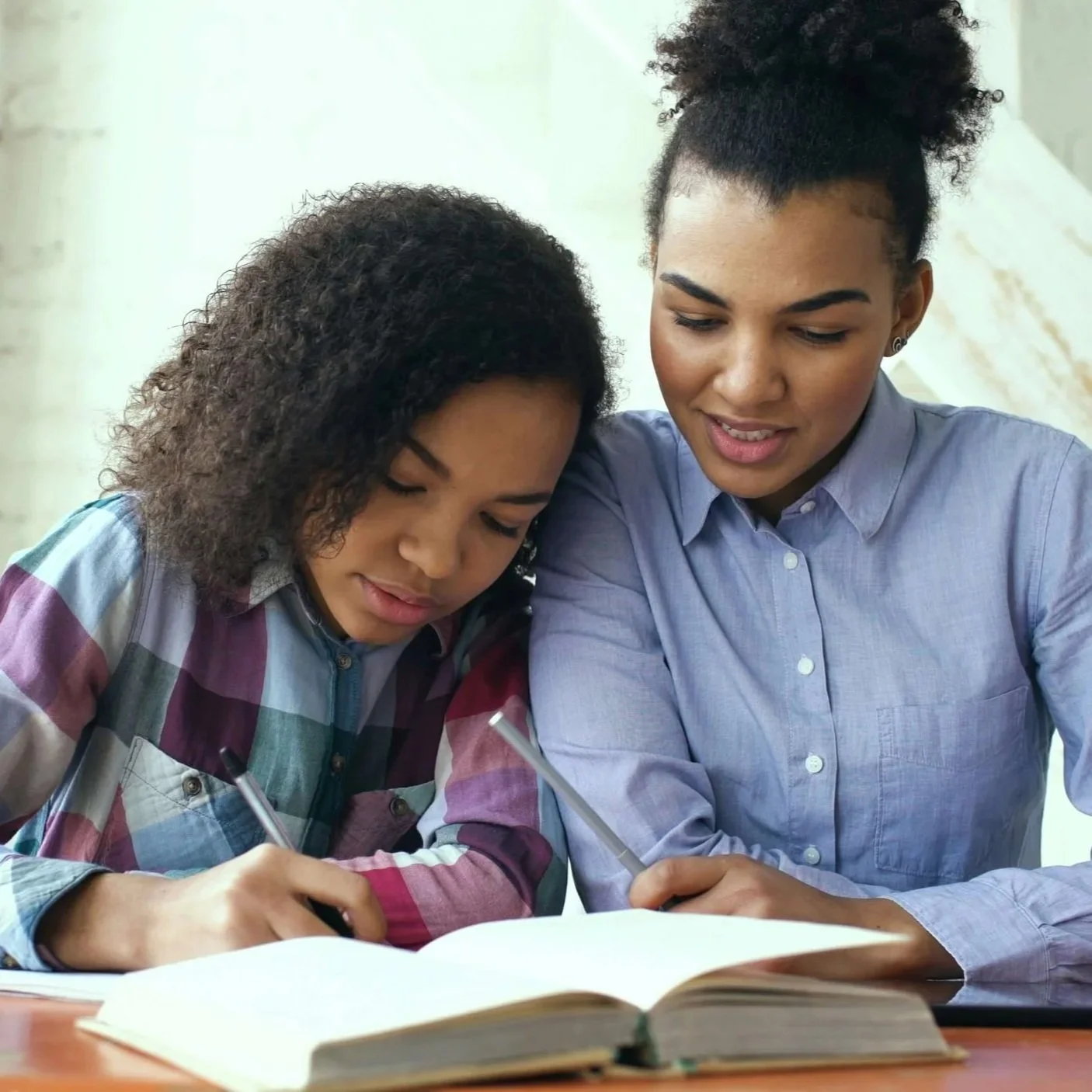 A young girl and a woman are sitting at a table, writing in a large book together. They are focused on their work, with the girl holding a pen and the woman watching her. Both are dressed casually, the girl in a colorful plaid shirt and the woman in a light blue button-up shirt.