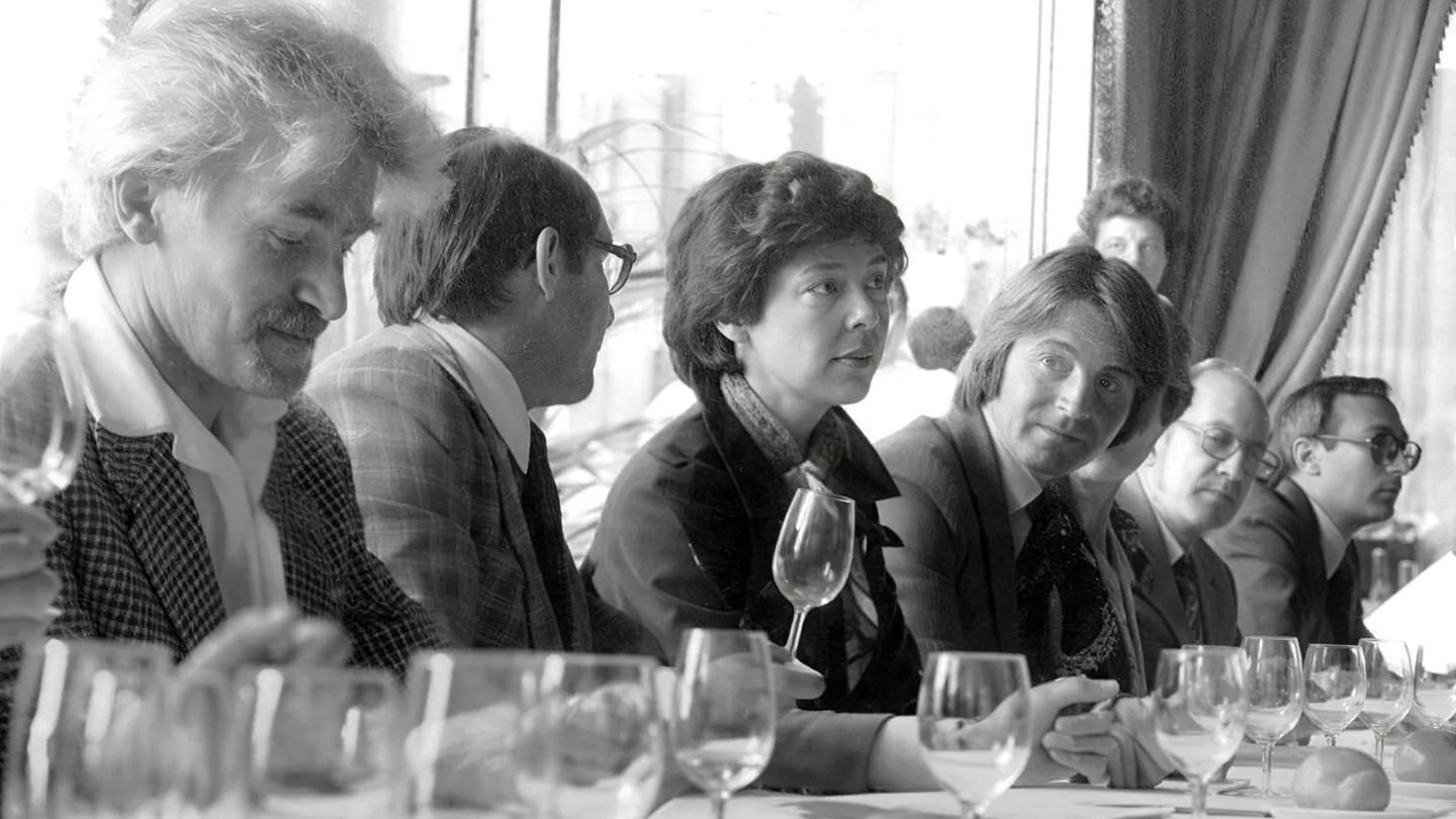 A moody 1970s editorial-style blind wine tasting table, with unlabeled bottles, partially filled wine glasses, handwritten scorecards and pencils, suggesting an elegant Paris hotel tasting in 1976.