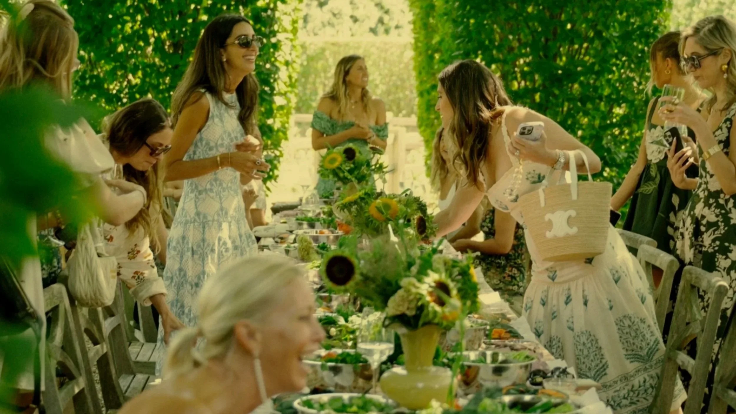 A group of women gathered around an outdoor dining table decorated with sunflowers and other flowers, enjoying a meal on a sunny day with lush green trees in the background.