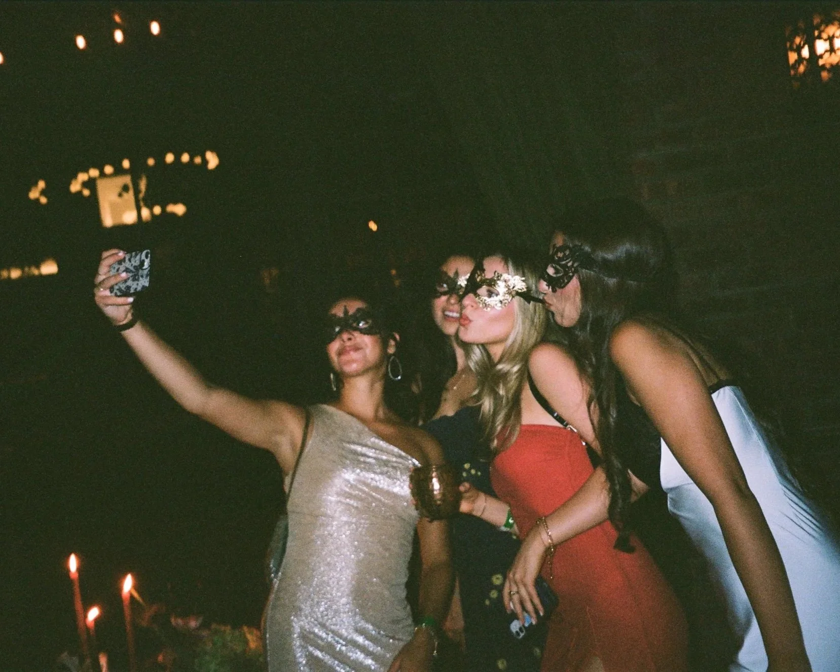 Group of women taking a selfie at a party or celebration, wearing festive masks and dresses, with blurred lights in the background.