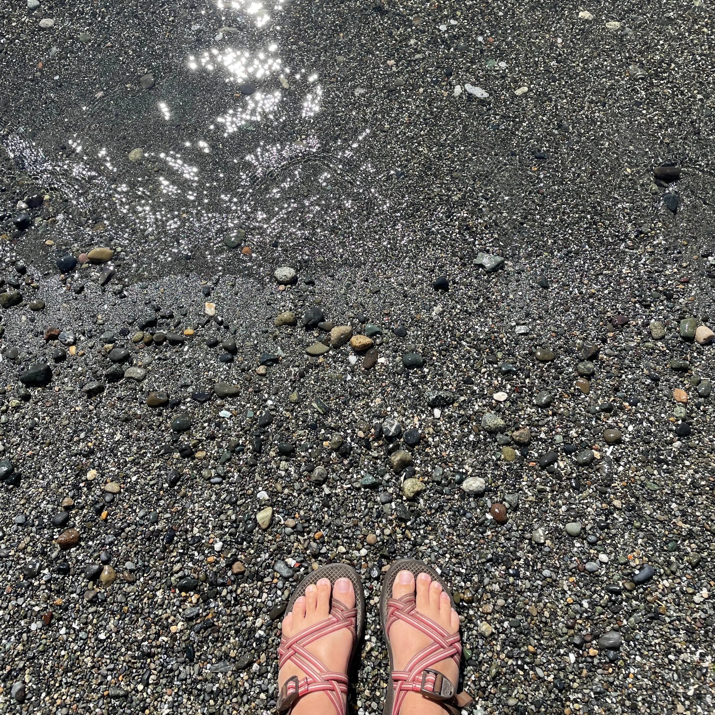 A picture of my feet in sandals standing a sandy beach at the edge of the water.