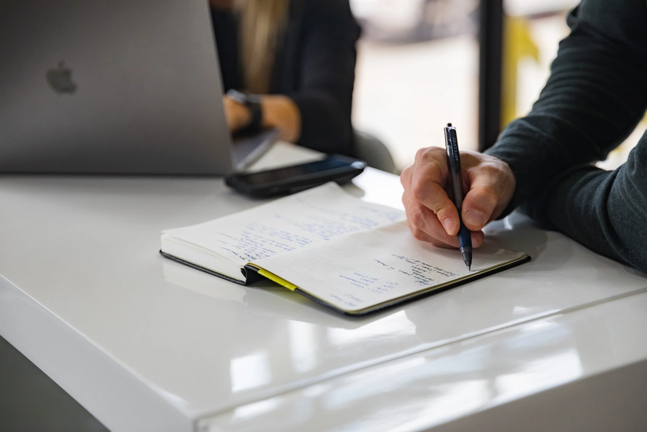 A person writing notes in a notebook with a pen on a white table, with a laptop and smartphone nearby.