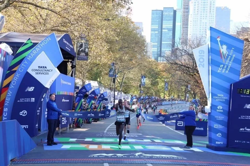 Finish line of the 2019 New York City Marathon with runners crossing, spectators, and city buildings in the background.
