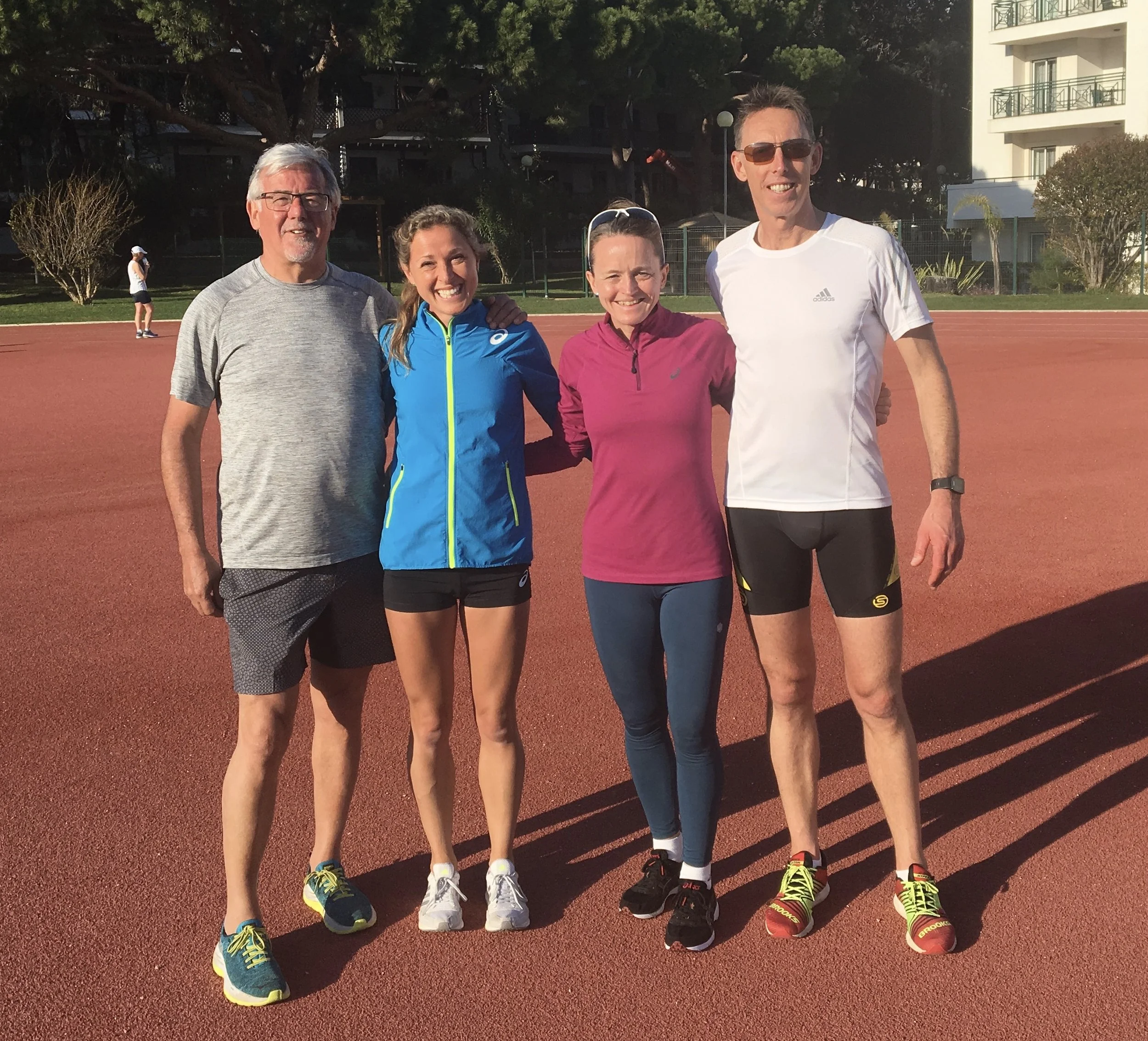 Four people standing together on a track, dressed in running attire, smiling at the camera in sunny weather.