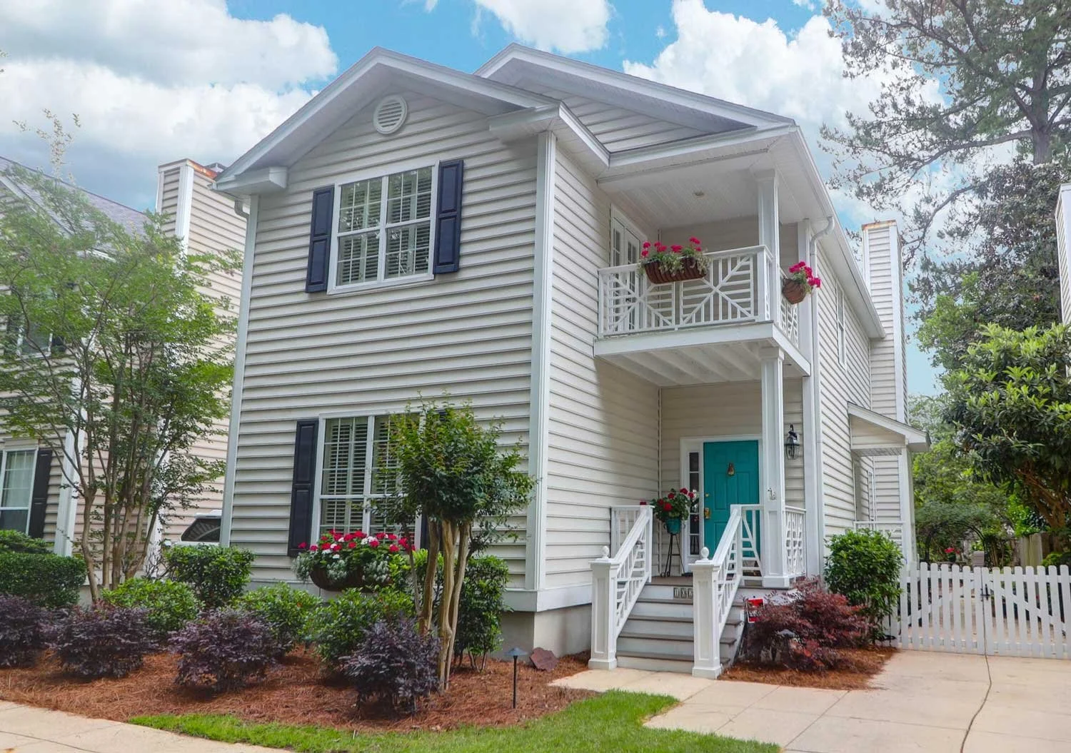 A two-story white house with black shutters, a blue front door, a small balcony with flower pots, and a landscaped yard with shrubs and trees.