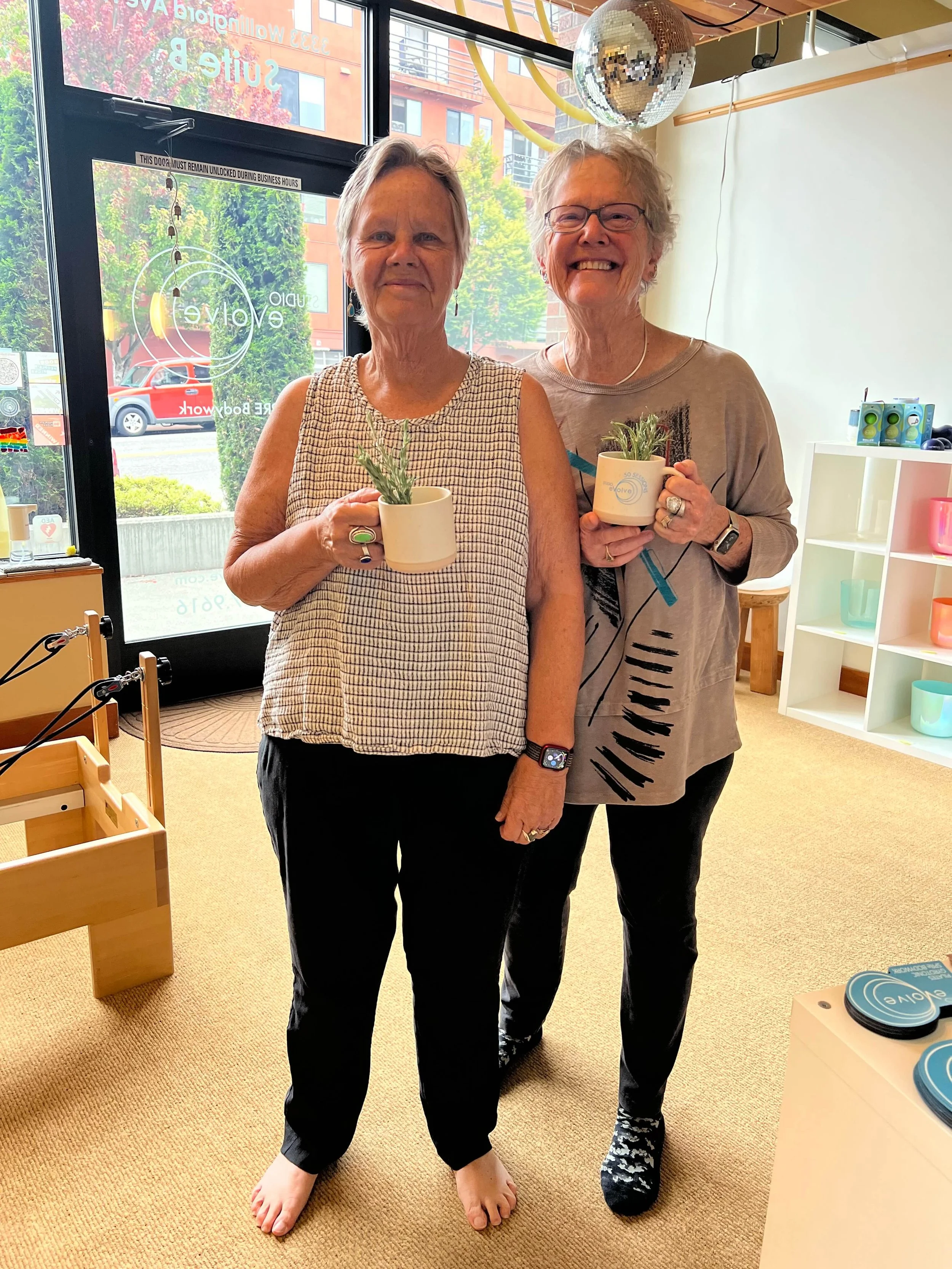 two women in their 70s smiling and holding studio evolve - Pilates studio mugs