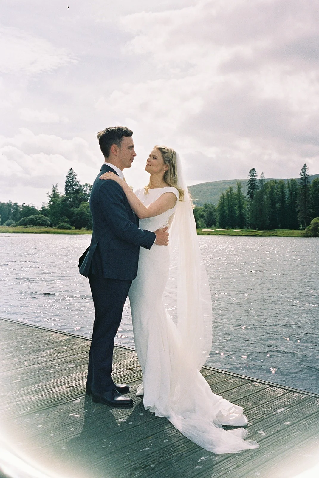 A bride and groom standing on a wooden dock by a river, gazing at each other with a scenic green landscape and cloudy sky in the background.
