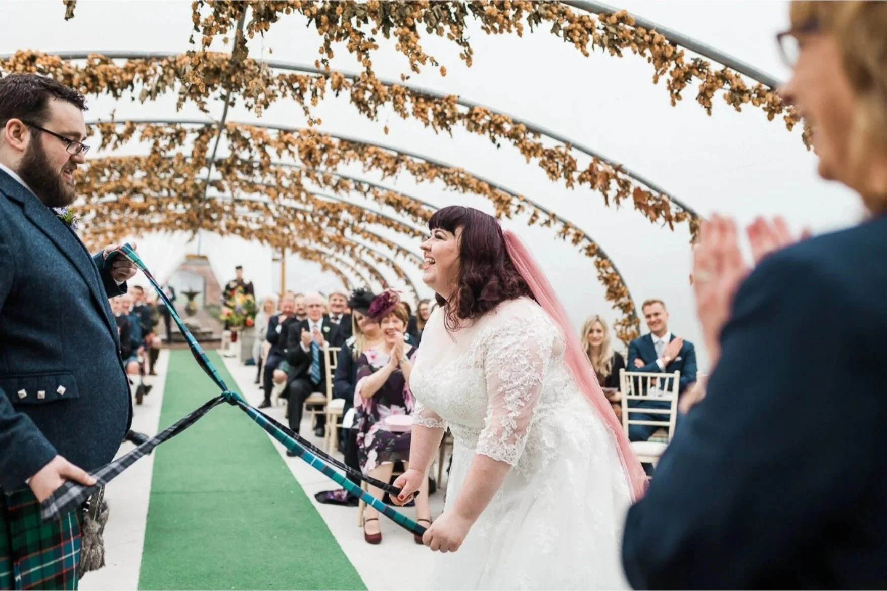 A bride and groom exchange vows in a wedding ceremony, holding a tartan cloth, with guests seated and clapping in the background under a floral arch.