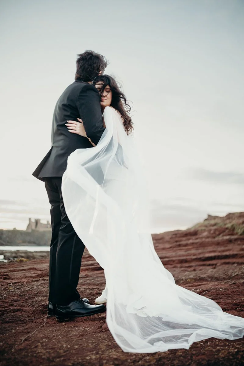 A couple in wedding attire embracing on a rocky landscape near the coast