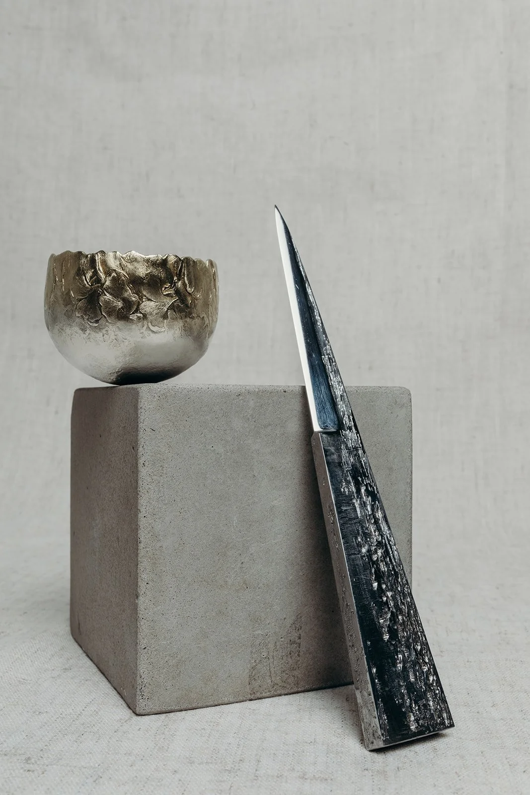 A sculpture display with a metallic bowl on a concrete pedestal and a black textured knife leaning against the pedestal.