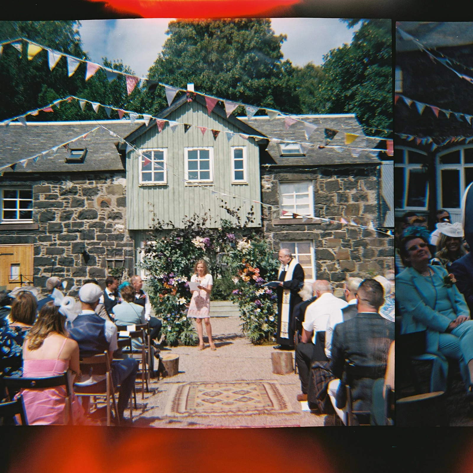 A wedding ceremony outdoors in front of a stone house decorated with bunting. The bride and officiant stand in front of floral arrangements, while guests sit on chairs watching.