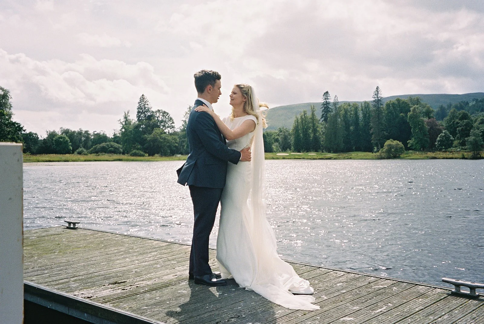 A bride and groom stand close together on a wooden dock by a lake, embracing and gazing into each other's eyes with a scenic background of trees, hills, and cloudy sky.