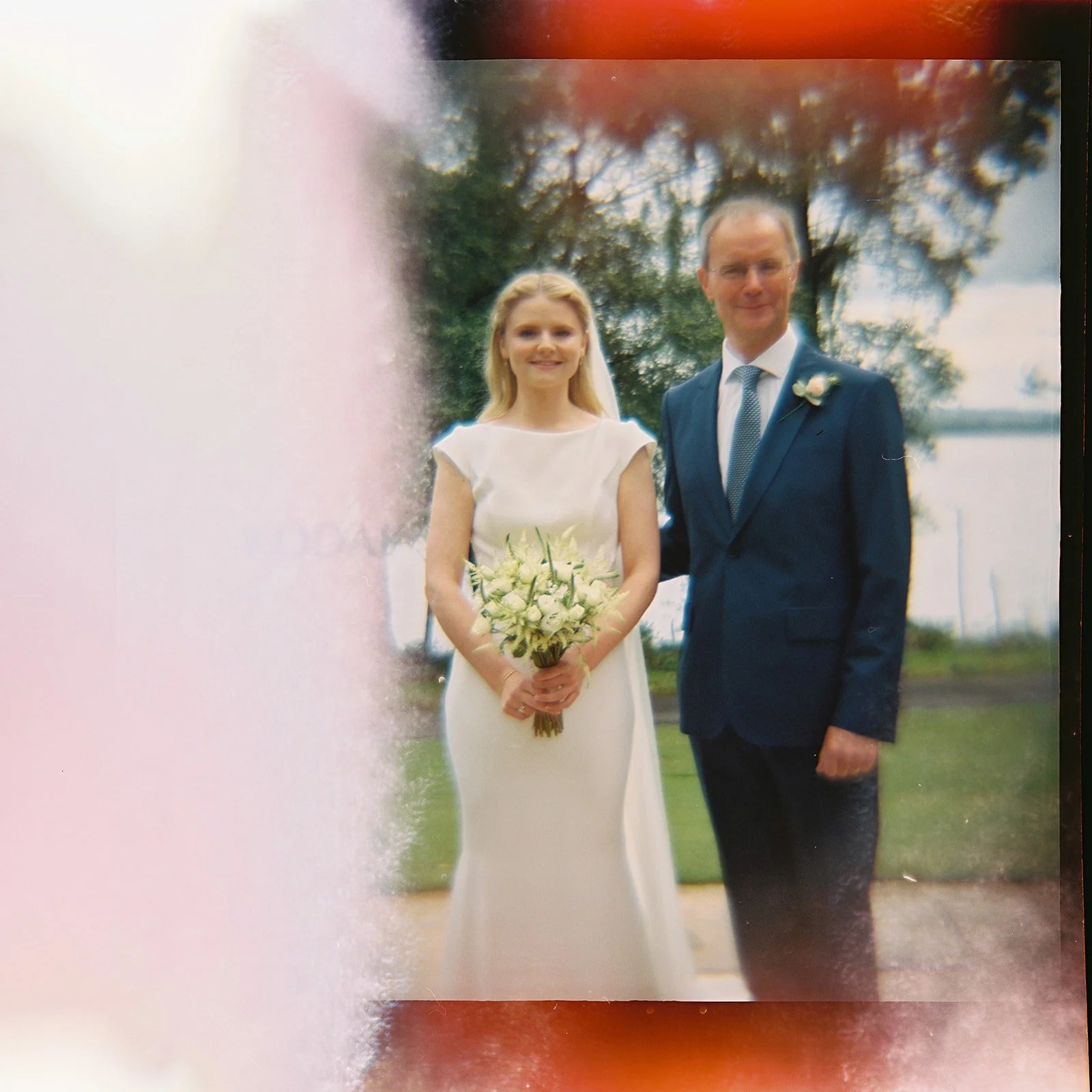 A bride and groom standing outdoors near a large tree with water in the background. The bride is holding a bouquet of white flowers, and the groom is wearing a dark suit with a boutonnière.