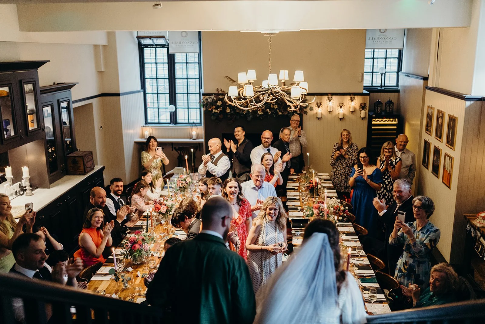 Wedding reception with bride and groom at the head of a decorated table, surrounded by seated and standing guests clapping and smiling in a warmly lit venue with elegant decor.
