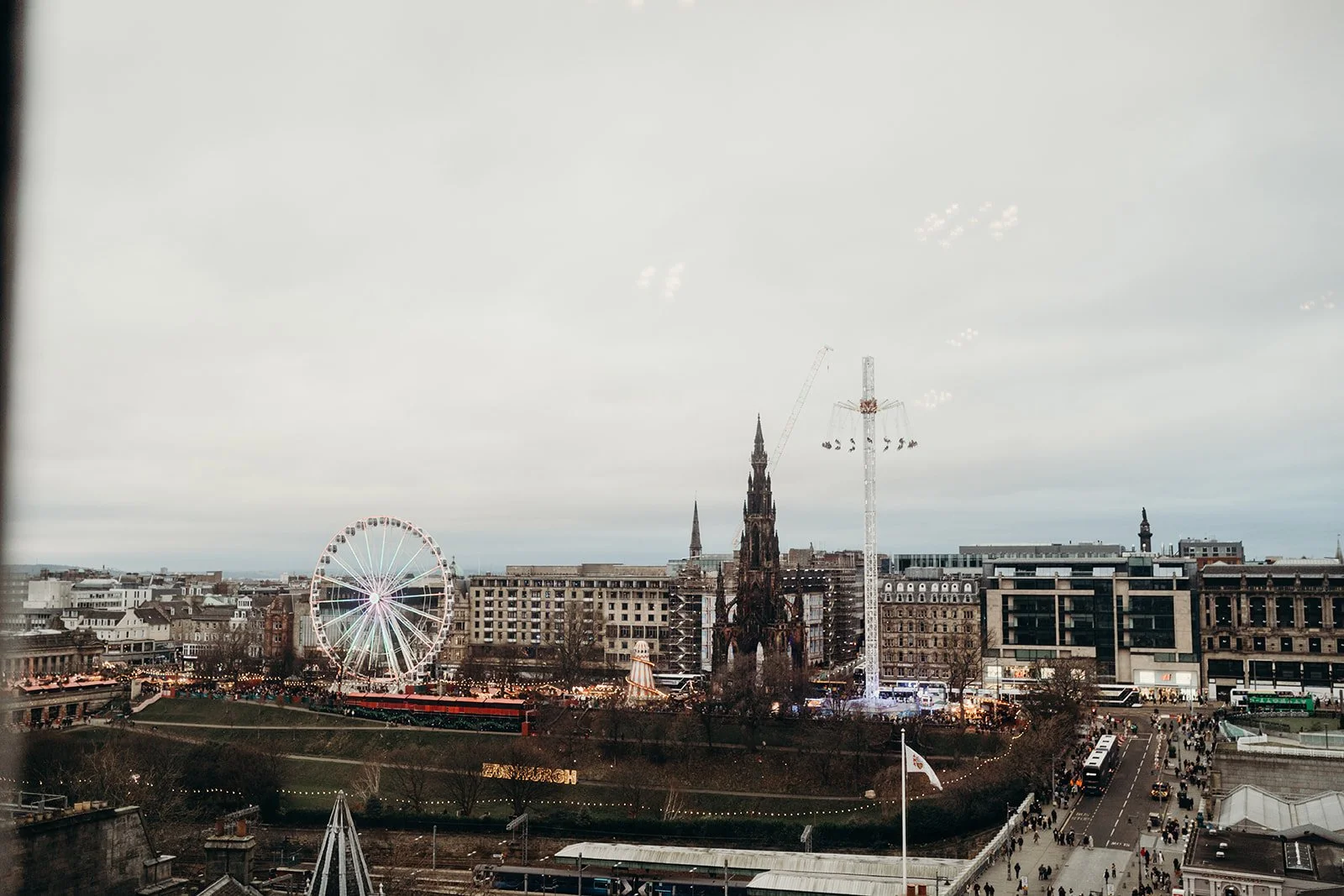 A cityscape with a Ferris wheel, a tall historic-looking building under construction, and an amusement park with rides and crowds, overcast sky.