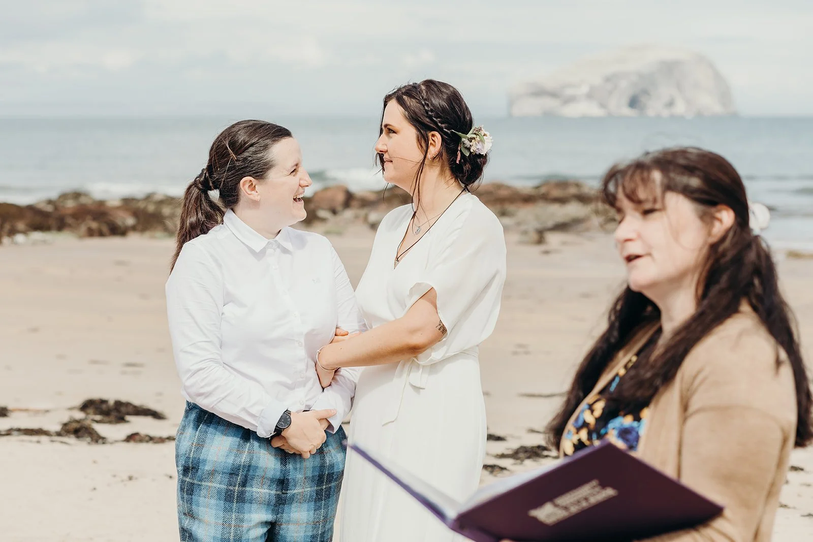 Two women exchanging vows at a beach wedding, with another woman holding a book or folder, and the ocean and rock formation in the background.